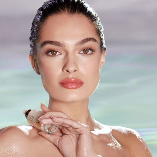 A light-tone model with glowy, glass skin, holding a luminous, ivory-coloured serum bottle with a dropper lid, while at the pool with her shoulders bare. 