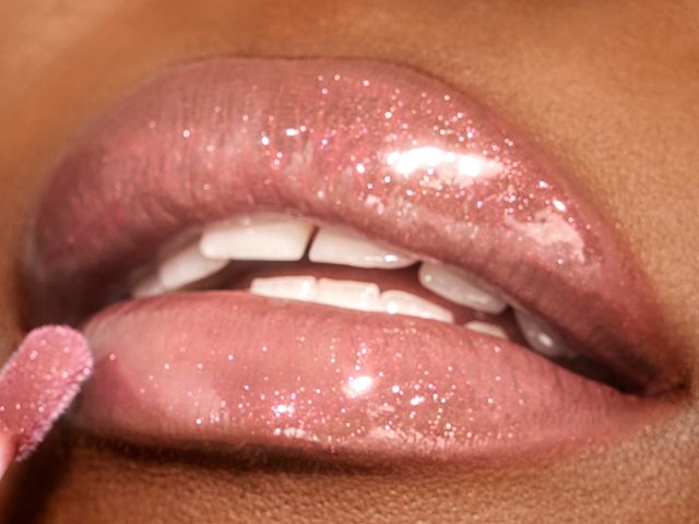 Close-up of a deep-tone model applying a shimmery, nude pink lip gloss.