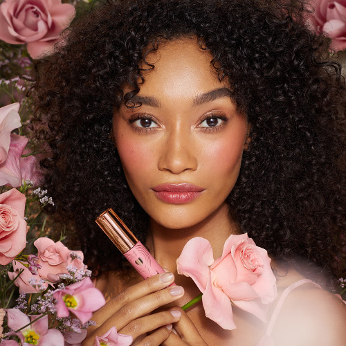 A deep-tone brunette model with brown eyes wearing a soft pink lip tint and holding the lip tint in one hand and a flower in the other.