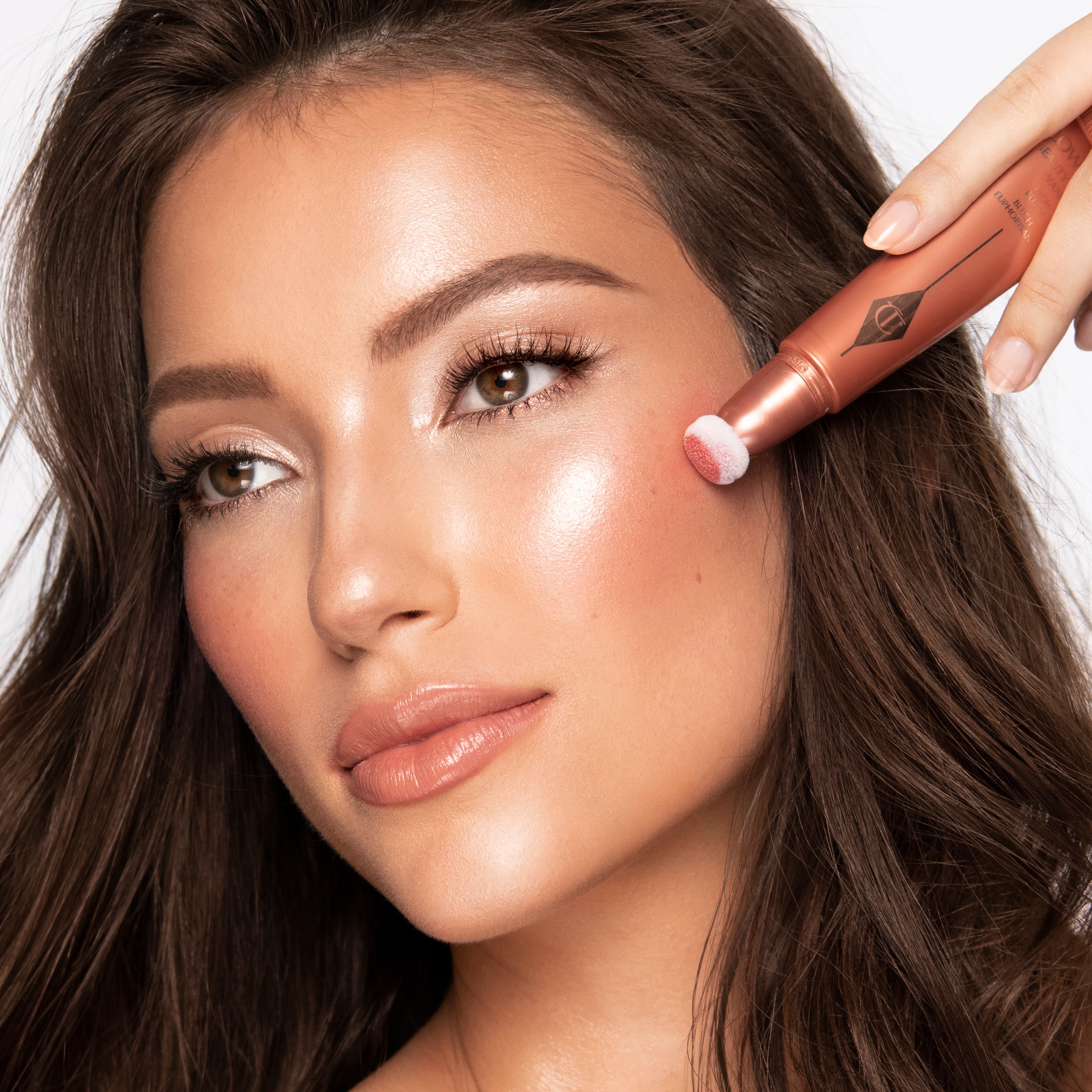 Medium-tone brunette model wearing glowy, nude coral-pink soft glam makeup look while getting applying a glowy coral blush applied from liquid highlighter blush wand.