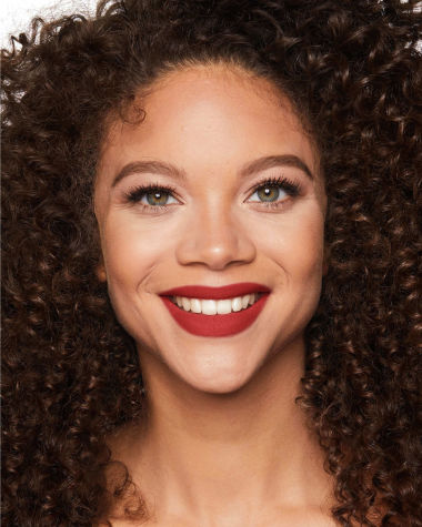 A medium-tone model with green eyes wearing a muted, matte brick red lipstick. 