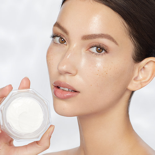 Fair-tone brunette model with glowy, flawless, glass skin, holding an open jar of pearly-white face cream.