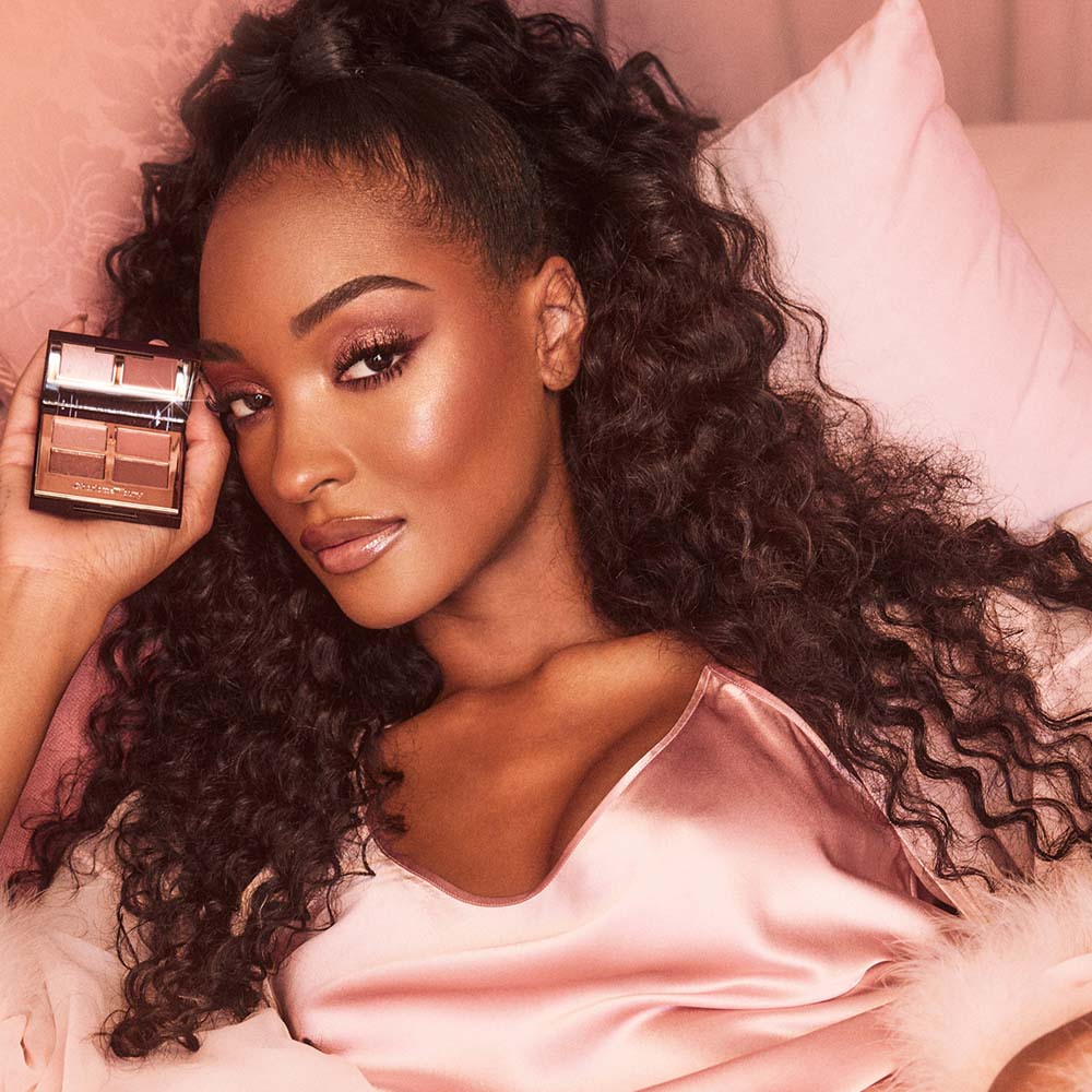 Deep-tone model with brown eyes wearing berry pink lipstick with a gloss on top with eye makeup in shades of pearlescent rose gold, dusky rose, berry brown and rose-bud pink while holding up a quad eyeshadow palette used to create the eye look.