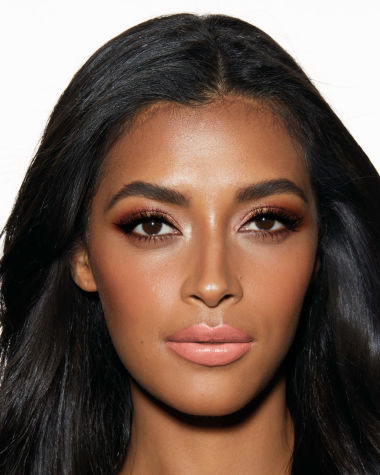 A deep-tone model with brown eyes wearing smokey brown and gold eye makeup with warm pink blush and glossy nude-pink lips