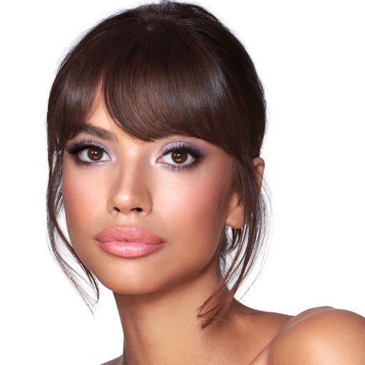 A medium-tone brunette model with brown eyes wearing shimmery lilac and champagne eye makeup with nude pink blush and coral lipstick. 