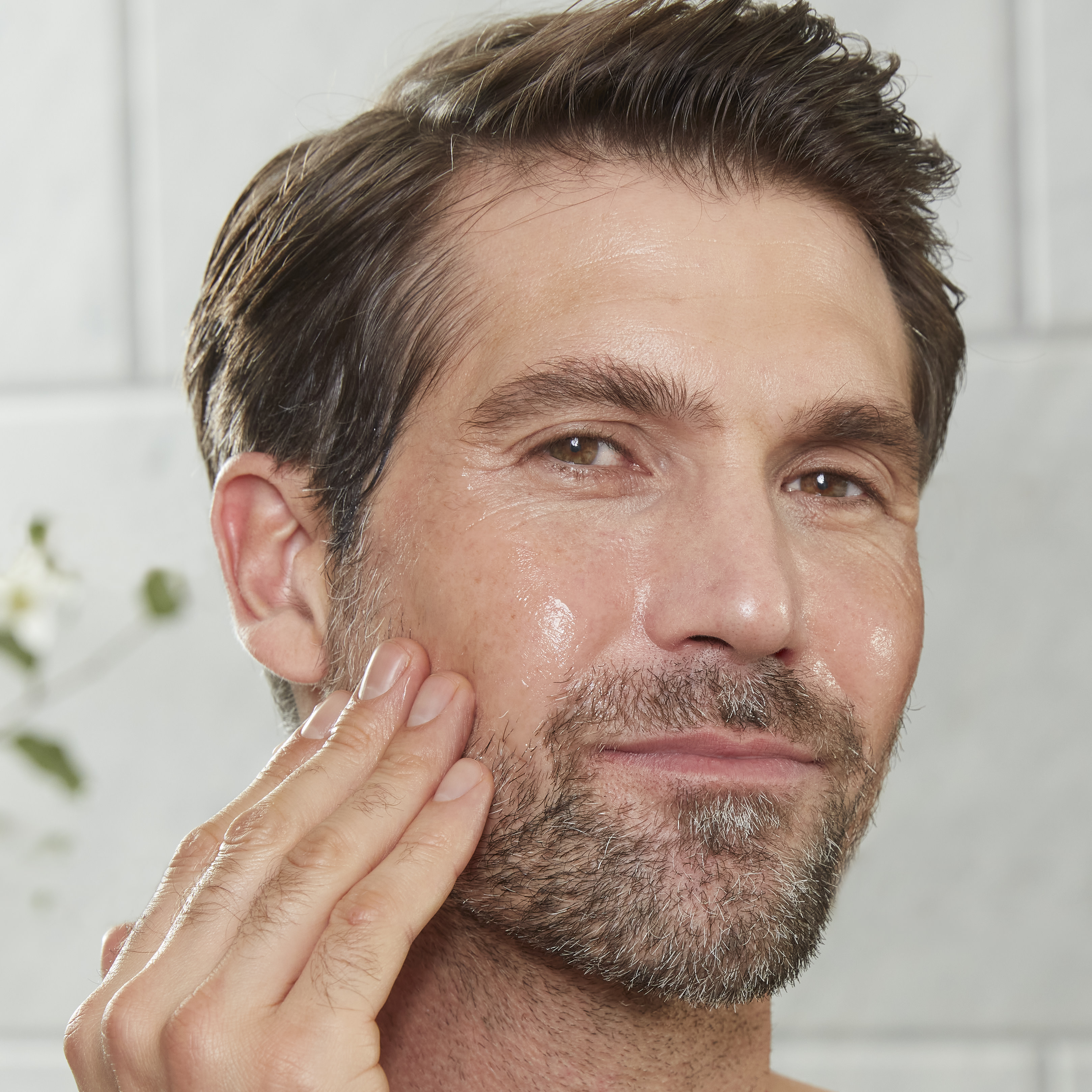 A light-tone male model with mature yet glowy, smooth, and radiant skin washing off an exfoliating mask while holding the white-coloured tube in his hand.