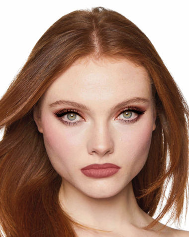 A fair-tone model with grey eyes wearing shimmery brown and gold eyeshadow with black kohl liner on her upper lid and lower waterline, a cool-toned glowy pink blush, and a berry-pink matte lipstick. 