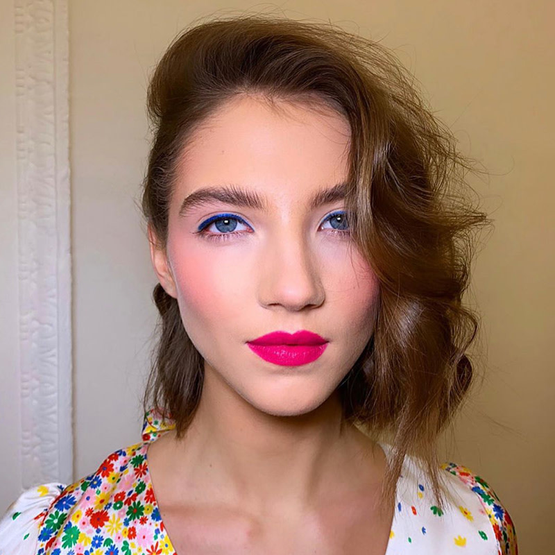Fair-toned brunette model with blue eyes wearing rosy make-up with red lipstick and wearing a white dress with multi-coloured flowers on it.