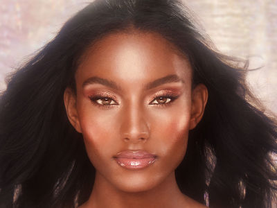 A deep-tone brunette model with an extremely glowy face base, rose-gold eye makeup, and nude pink glossy lips. 