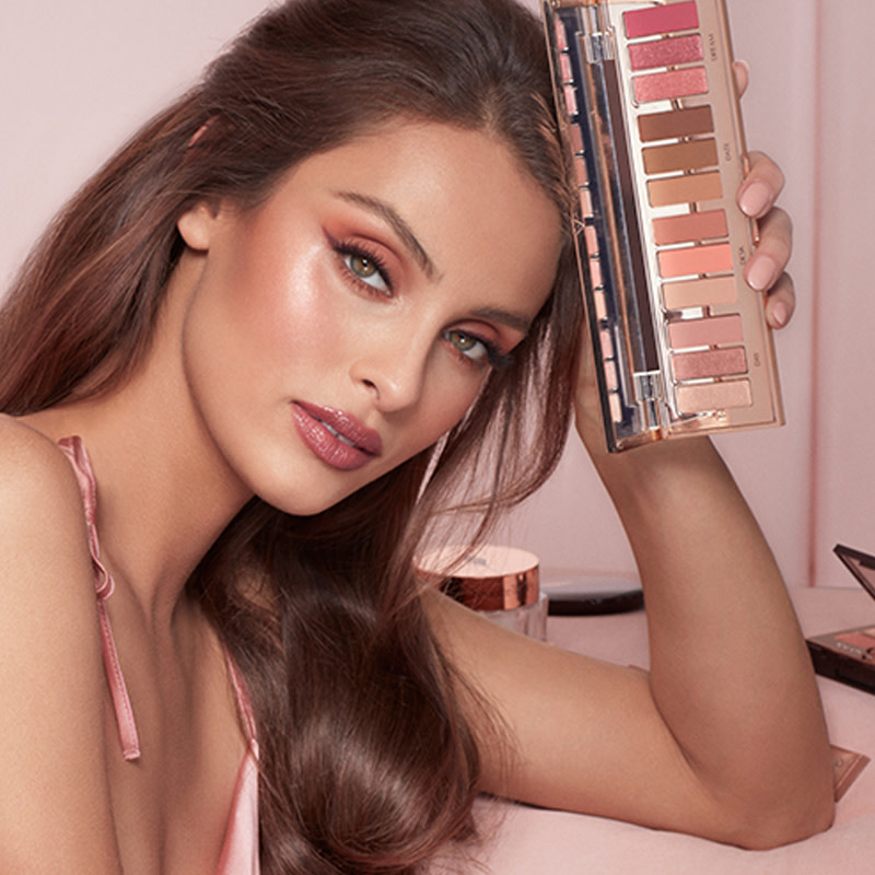 A medium-tone brunette model wearing glowy, berry-pink and nude pink makeup while holding an open, eyeshadow palette in matte and shimmery nude shades. 