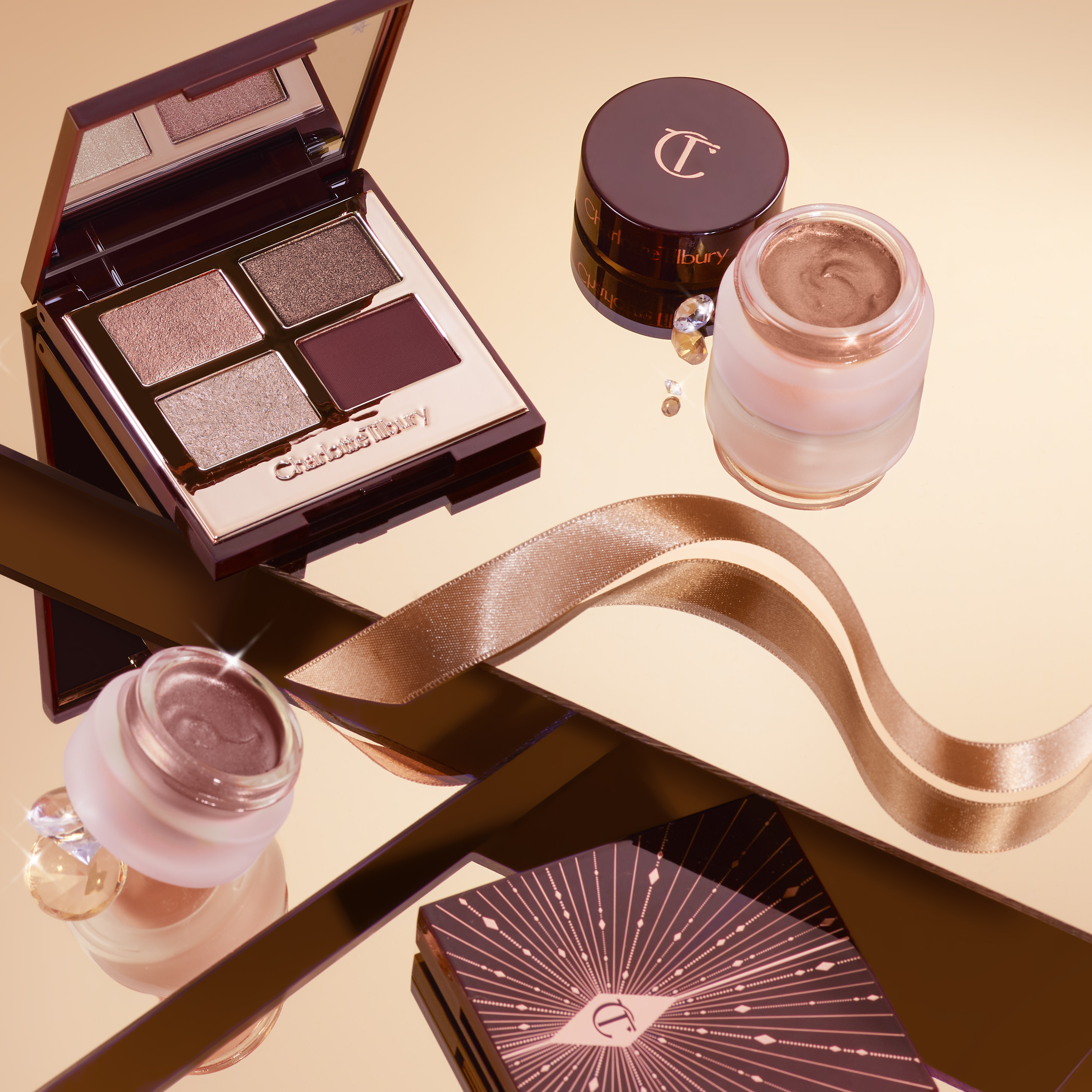 Banner with an open, mirrored-lid quad eyeshadow palette with red, brown, rose gold, and beige-gold-coloured eyeshadows surrounded by cream eyeshadows in petite glass pots and golden ribbons. 