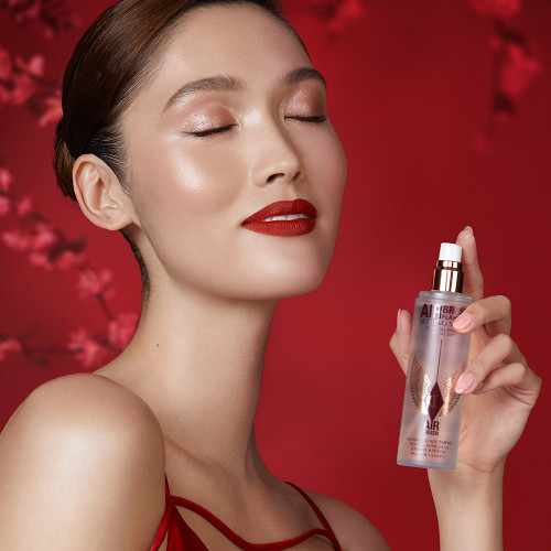 A fair-tone brunette model wearing dewy nude makeup with bold red lipstick while applying a setting spray. 