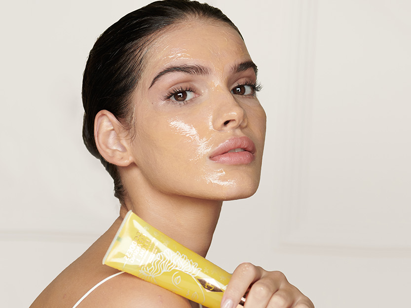 A light-tone model with flawless skin holding a lemon-yellow-coloured facial cleanser with it also applied on her face. 