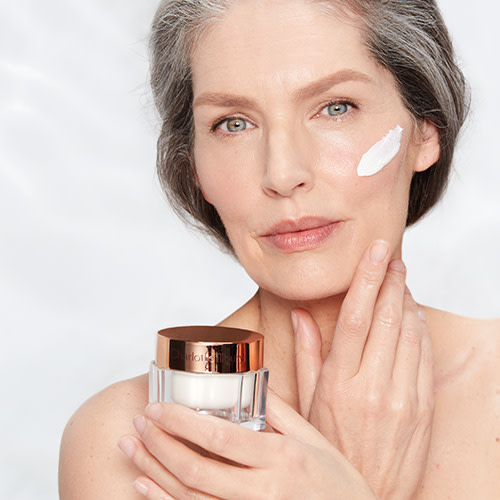 Fair-tone model with glowy, flawless, mature skin, and holding a closed jar of pearly-white face cream with a gold-coloured lid.