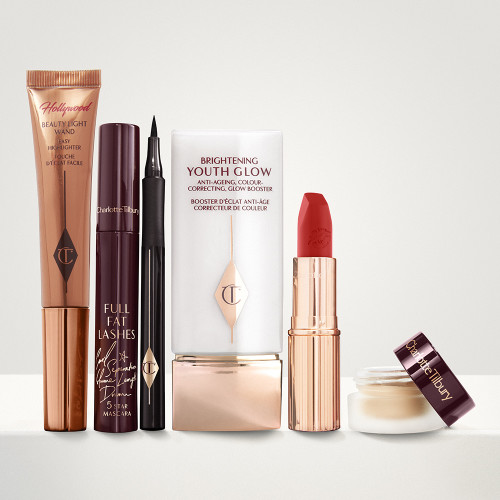 A copper-coloured highlighter wand, mascara in dark crimson packaging, an open eyeliner pen, a primer in white and gold packaging, an open lipstick in a bright orange-red shade, and cream eyeshadow in an open glass pot. 