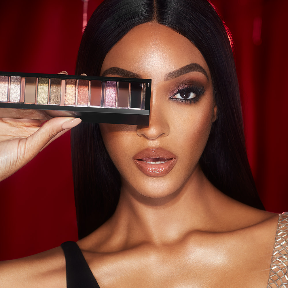 Deep-tone model with brown eyes wearing smokey brown eye makeup with nude pink lips and holding up an open, 12-pan eyeshadow palette with matte and shimmery shades in beige, pink, gold, green, brown, peach, and black.