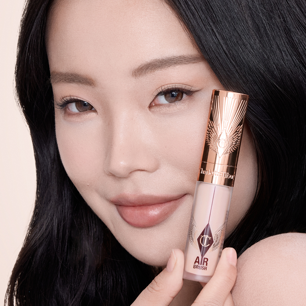 Close-up of a hand holding a rose-gold tube of Charlotte Tilbury Airbrush Flawless Foundation, with the product name partially visible. The background features smooth, dark hair cascading down, creating a sleek and elegant contrast.