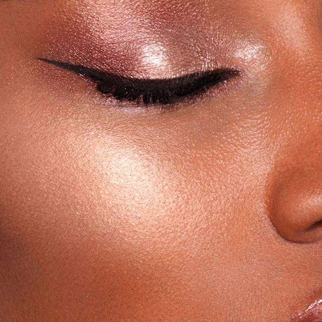 Hollywood Superstar Glow Highlighter on deep-tone model close-up Close-up of a deep-tone model wearing shimmery rose gold eyeshadow with black eyeliner in a sharp wing and powder highlighter in a glowing, soft, rose gold shade.