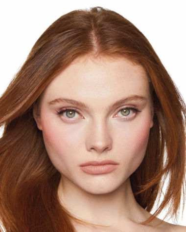 A fair-skin, brunette model wearing a warm rosy-pink blush with a nude lipstick. 