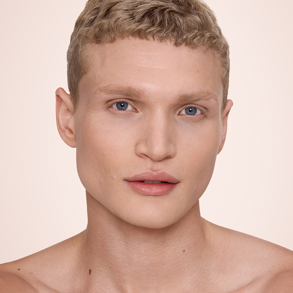 Close-up of a hand holding a rose-gold tube of Charlotte Tilbury Airbrush Flawless Foundation, with the product name partially visible. The background features smooth, dark hair cascading down, creating a sleek and elegant contrast.
