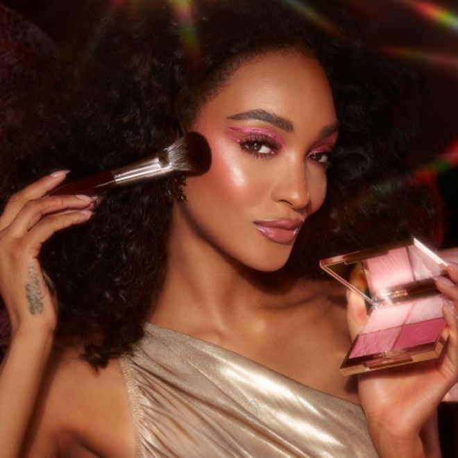 Pillow Talk Beautifying Face Palette on deep-tone model Deep-tone brunette model with brown eyes wearing full-glam, extremely glowy pink makeup while holding an open, face palette with matte and shimmery eyeshadows, blushes and highlighters in shades of pink and gold with a mirrored lid.