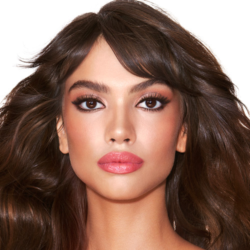 A medium-tone model with brown eyes wearing soft fawn and brown eye makeup with glowy terracotta blush, and sheer, glossy rose-pink lips.