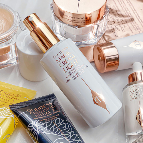 A collection of skincare that includes an open, face cream in a white-coloured bottle with a gold-coloured pump dispenser, luminous ivory-coloured serum in a glass bottle, charcoal facial scrub and lemon oil cleanser, day and night creams in glass jars with gold-coloured lids, and clay mask in a white tube with gold-coloured lid.