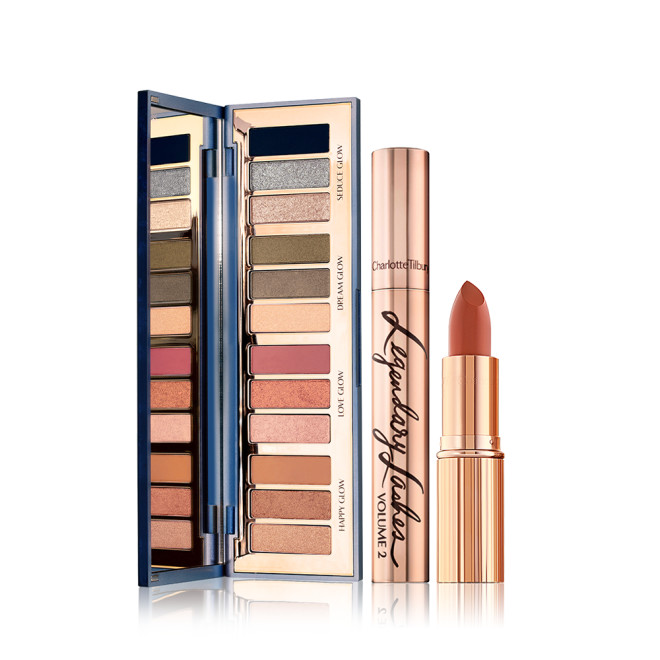 An open, mirrored-lid eyeshadow palette with eyeshadows in shades of pink, brown, peach, golden, blue, and green, along with a mascara with gold packaging, and an open lipstick in a medium-dark coral shade.
