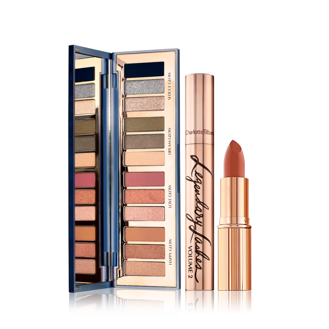 An open, mirrored-lid eyeshadow palette with eyeshadows in shades of pink, brown, peach, golden, blue, and green, along with a mascara with gold packaging, and an open lipstick in a medium-dark coral shade.