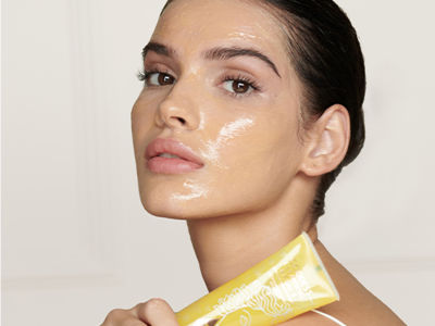 Light-toned brunette model holding a cleanser in a yellow tube with the cleanser applied to her face.