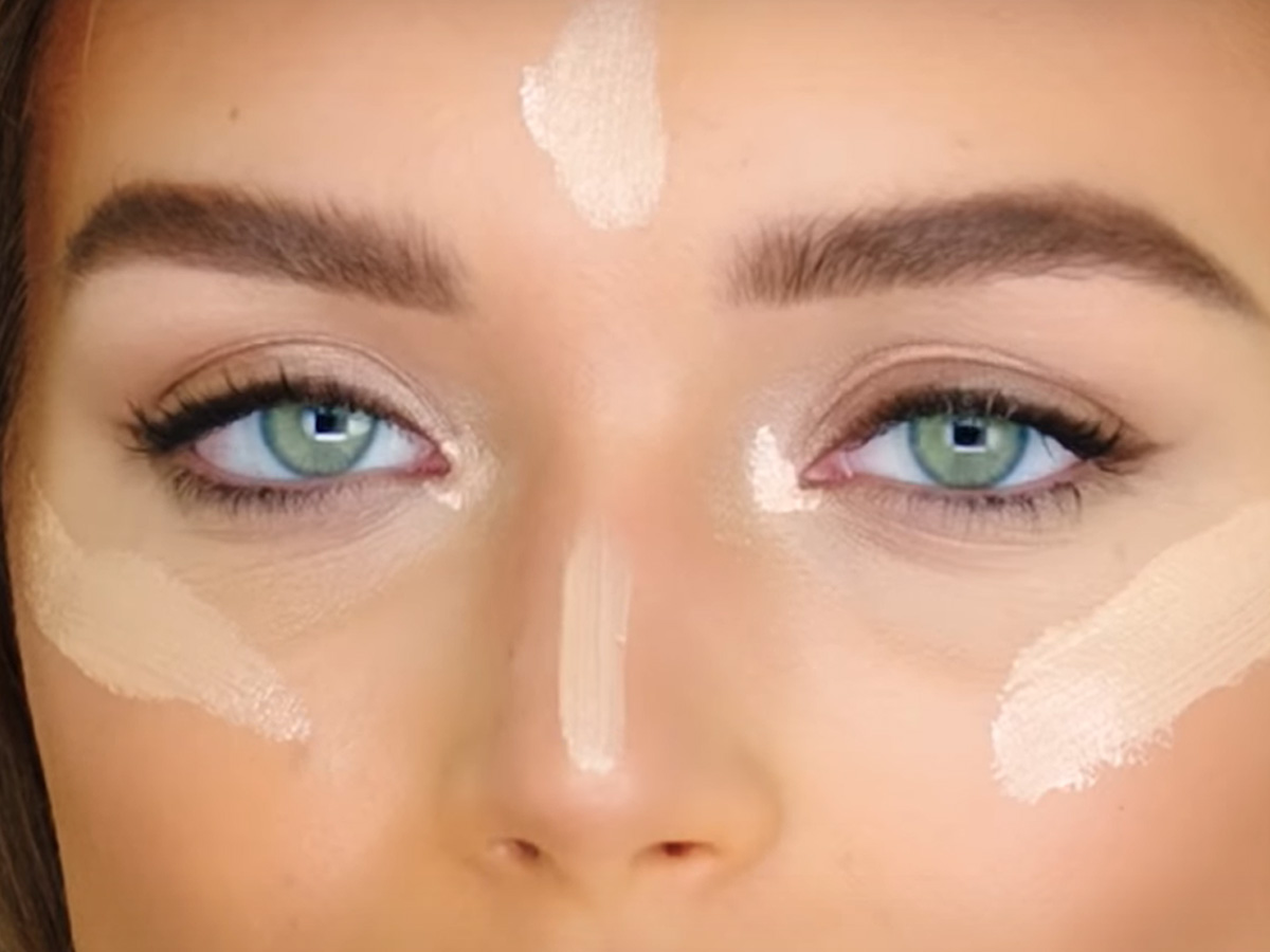 Close-up of a light-tone model with green eyes with champagne-coloured highlighter applied to forehead, nose, and cheeks but not blended yet. 