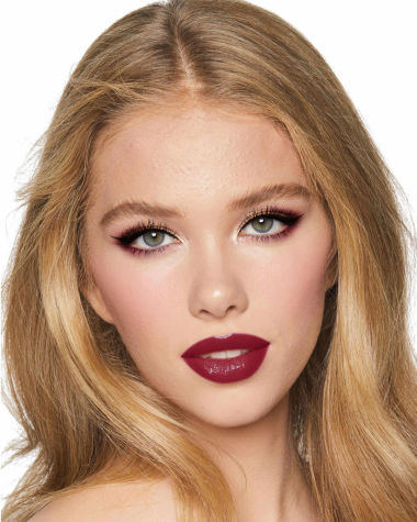 A fair-tone model with grey eyes wearing reddish-plum eye makeup, glowy face base, and a vampy-red lipstick with a satin-finish. 