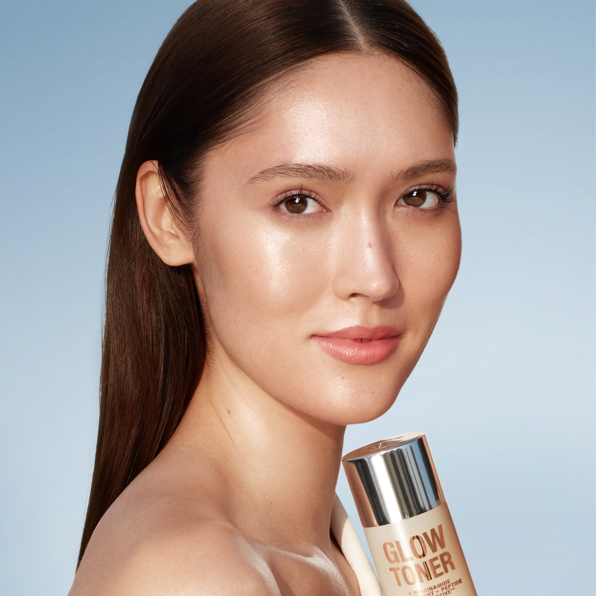 Fair-tone model with glowing, fresh skin holding a large toner bottle filled with a creamy, silvery-white-coloured toner with a silver-coloured lid. 