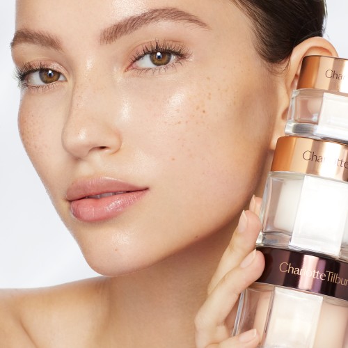 Fair-tone brunette model with glowy, glass skin holding pearly-white face cream, fawn-coloured eye cream, and peach-coloured night cream in glass jars with sleek lids.