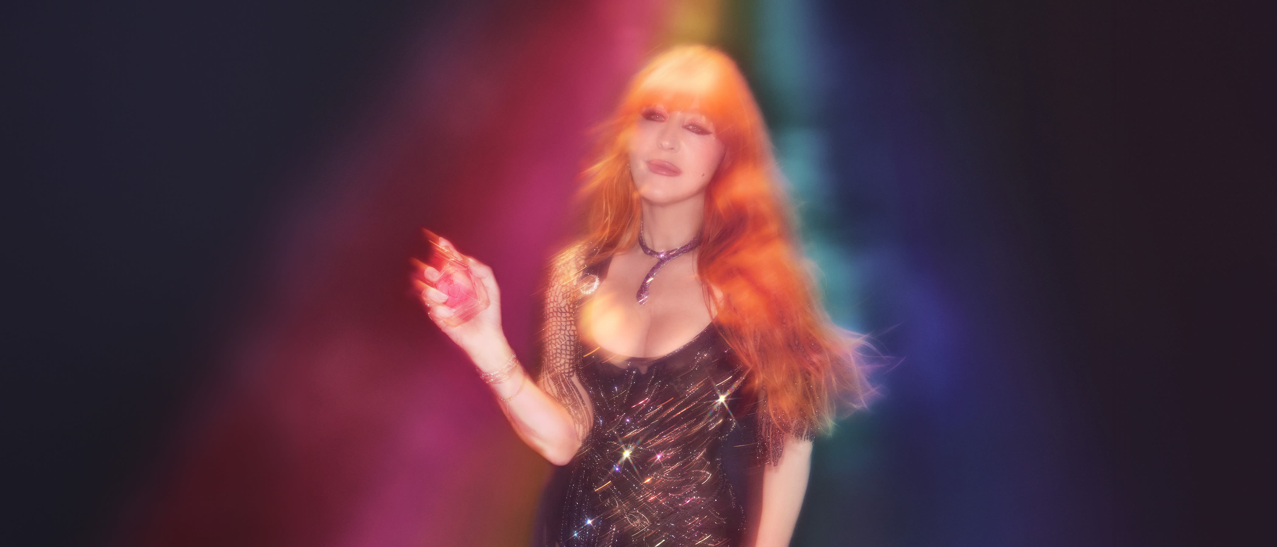 Charlotte Tilbury in a rainbow light wearing a sparkly black dress holding a bottle of perfume