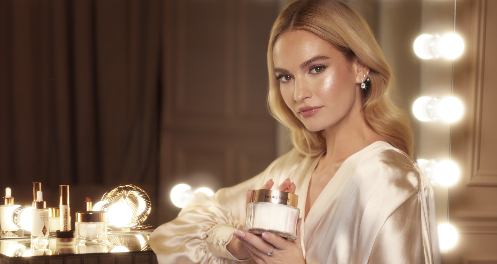 Banner with actress, Lily James, with glowy, glass skin, holding an open jar of pearly-white face cream.