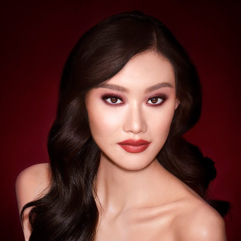 A fair-tone model with brown eyes wearing reddish-plum eye makeup, glowy face base, and a vampy-red lipstick with a satin-finish. 