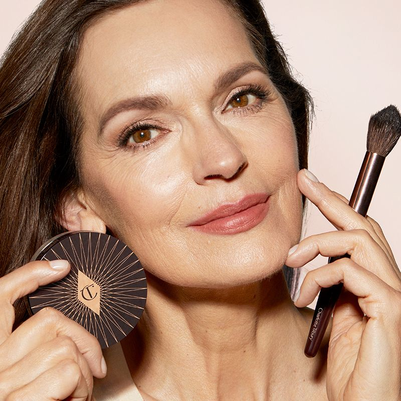 A light-tone model with mature skin wearing coral-red lipstick and eyeliner with a flawless, luminous base while holding a powder brush and holding Charlotte's Genius Magic Powder.