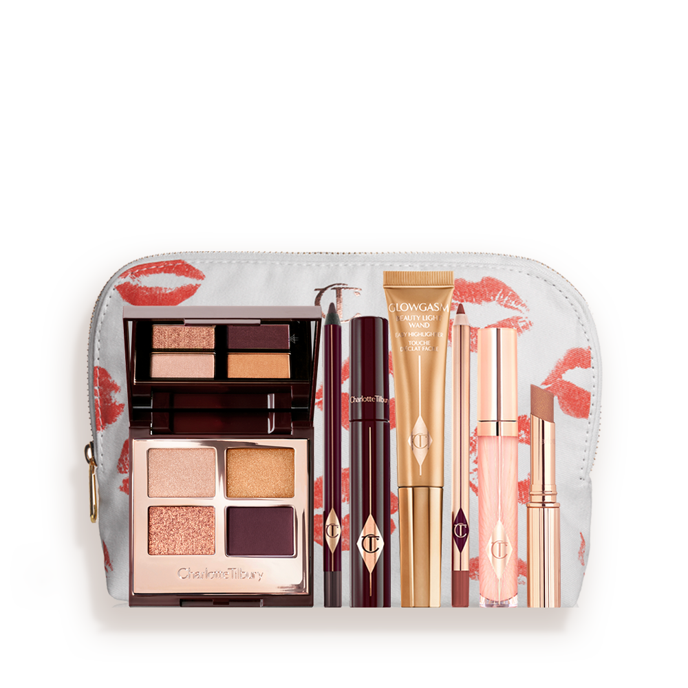 A highlighter wand in a honey-gold-coloured tube with a mascara, eyeliner pencil, quad eyeshadow palette with shimmery and matte rose gold, plum, and golden shades, an open lipstick in nude brown, lip liner pencil in soft plum, and a lip gloss in nude pink, and a white-coloured makeup pouch. 