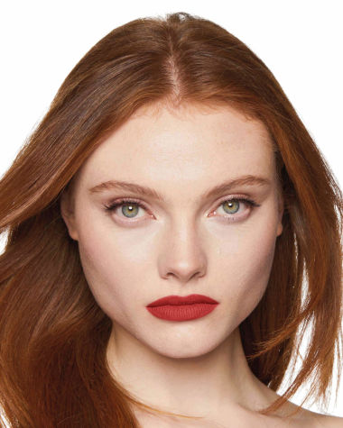 A fair-tone model with grey eyes wearing a muted, matte brick red lipstick. 