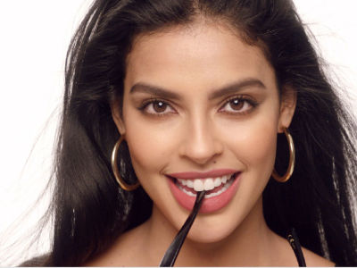 A medium-tone brunette model with brown eyes wearing rose-pink lipstick, smokey brown eyeshadow, with a matte-finish base, and large gold hoop earrings. 