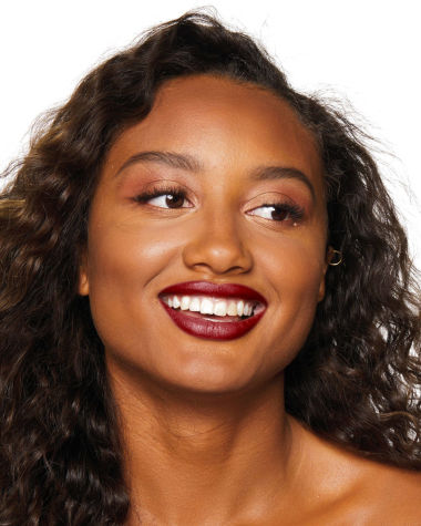 Deep-tone model with brown eyes wearing a muted wine-red lipstick with a satin finish.