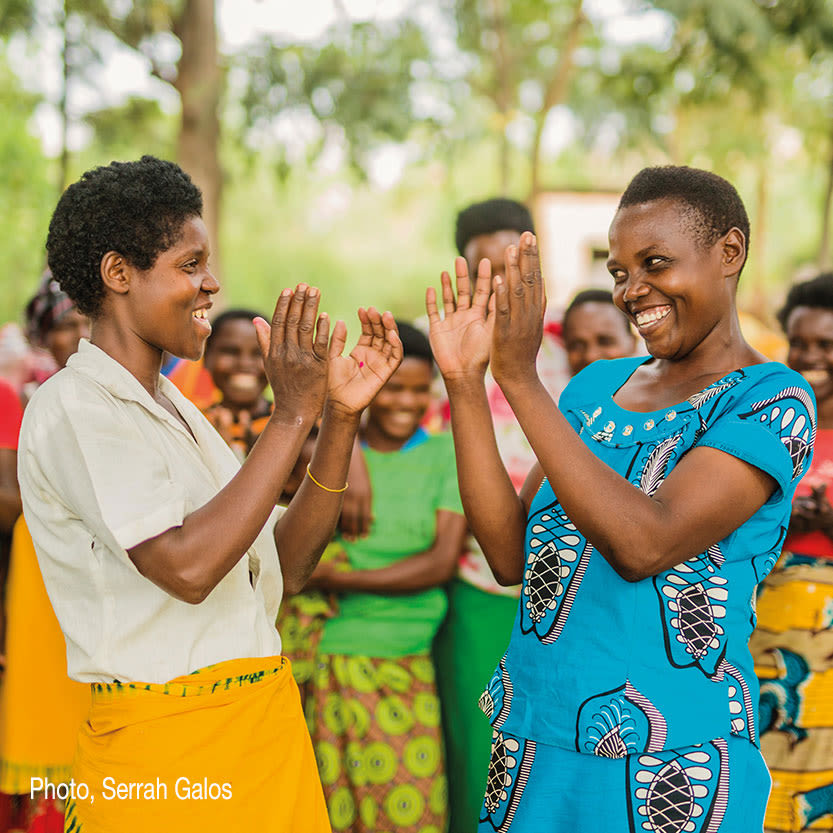 Two deep-tone women laugh and play in front of a large group of women laughing and enjoying themselves. 