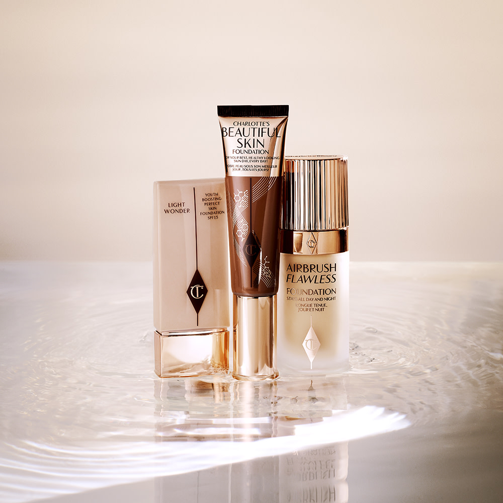 Foundation in a clear plastic bottle with a sleek gold coloured lid, foundation wand with gold packaging and a dark brown body that's the colour of the foundation inside along with a gold-coloured lid, and foundation in a frosted glass bottle with a gold-coloured lid.