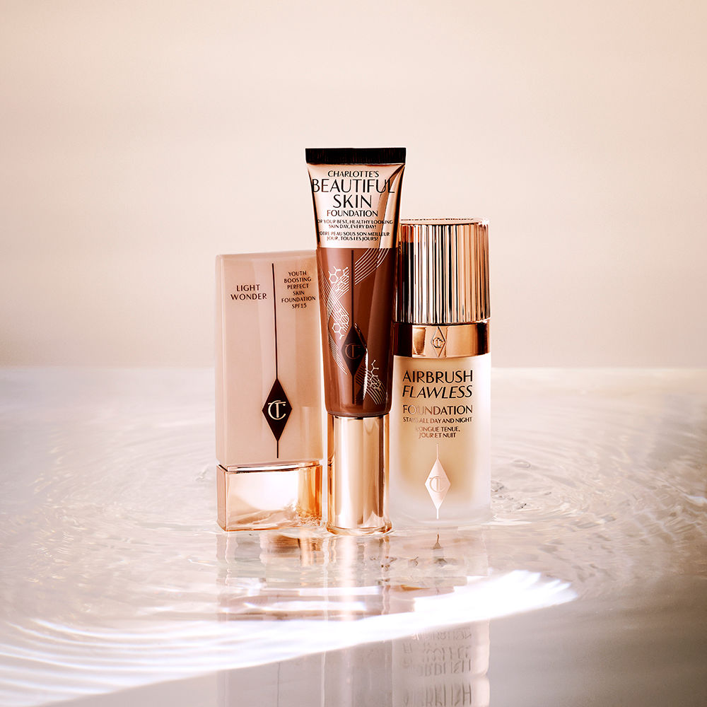 Foundation in a clear plastic bottle with a sleek gold coloured lid, foundation wand with gold packaging and a dark brown body that's the colour of the foundation inside along with a gold-coloured lid, and foundation in a frosted glass bottle with a gold-coloured lid.