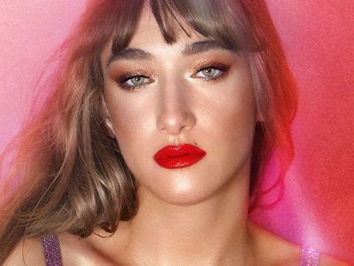 A medium-tone model with light hair wearing a bold red lip shade, shimmery bronze eyeshadow, and an extremely glowy face base. 