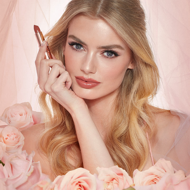 Fair-tone model with blue eyes wearing a universally flattering, peachy-nude lipstick with a satin-finish while holding the lipstick.