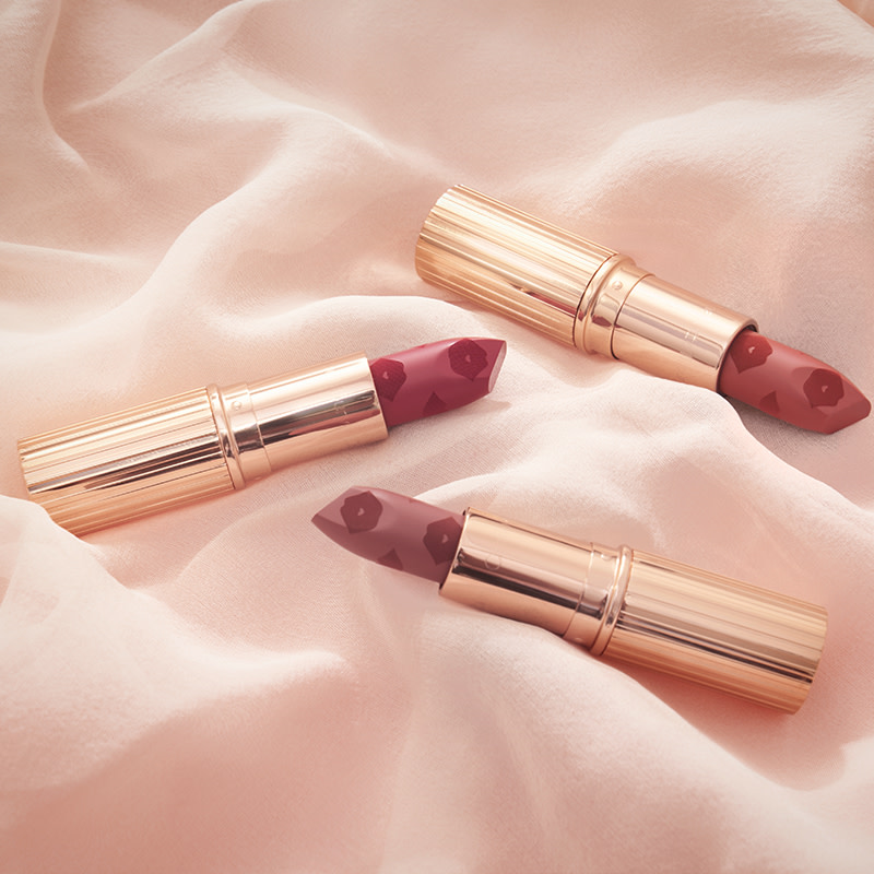 Three open lipsticks in shades of red, mauve, and terracotta with kiss prints all over, in golden-coloured tubes, placed on a champagne-coloured sheet. 