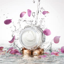 Thick and luscious, pearly-white face cream in a glass jar with sparkling water and rose petals falling on it. 