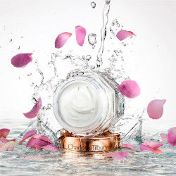 Thick and luscious, pearly-white face cream in a glass jar with sparkling water and rose petals falling on it. 